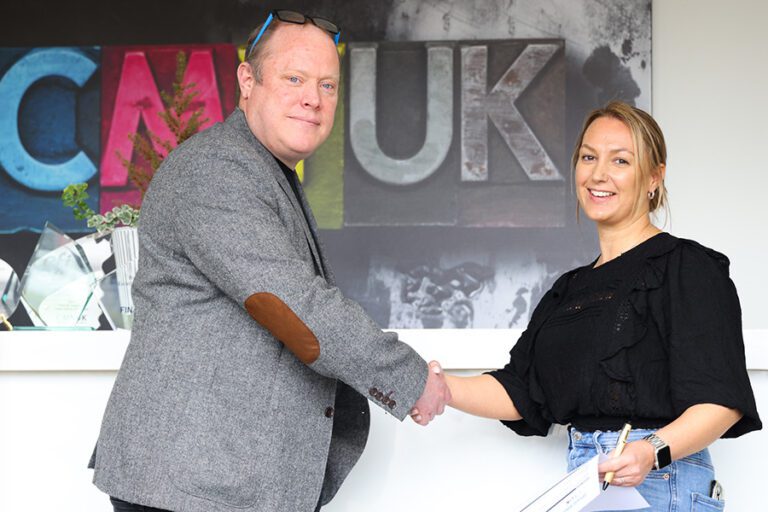 Chris Davies, event director of The Print Show wearing a grey tweed blazer with brown leather elbow pads on the left shaking hands with Sarah Neate, group marketing director of CMYUK standing in front of a colourful wall featuring the letters CMYUK